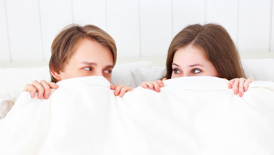 Casal se olhando, deitados na cama, dentro dos lençóis