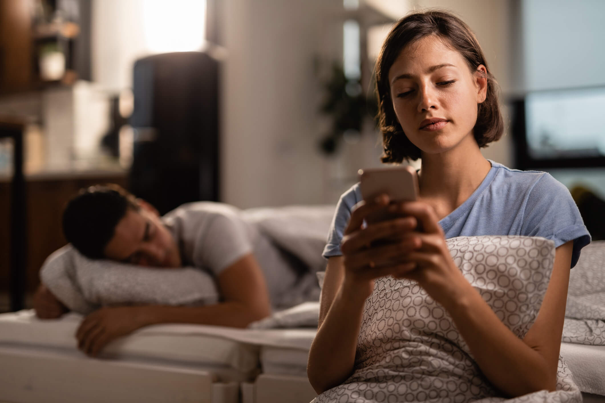 foto de homem dormindo ao fundo e mulher em primeiro plano usando o celular. imagem para representar sonhar com traição