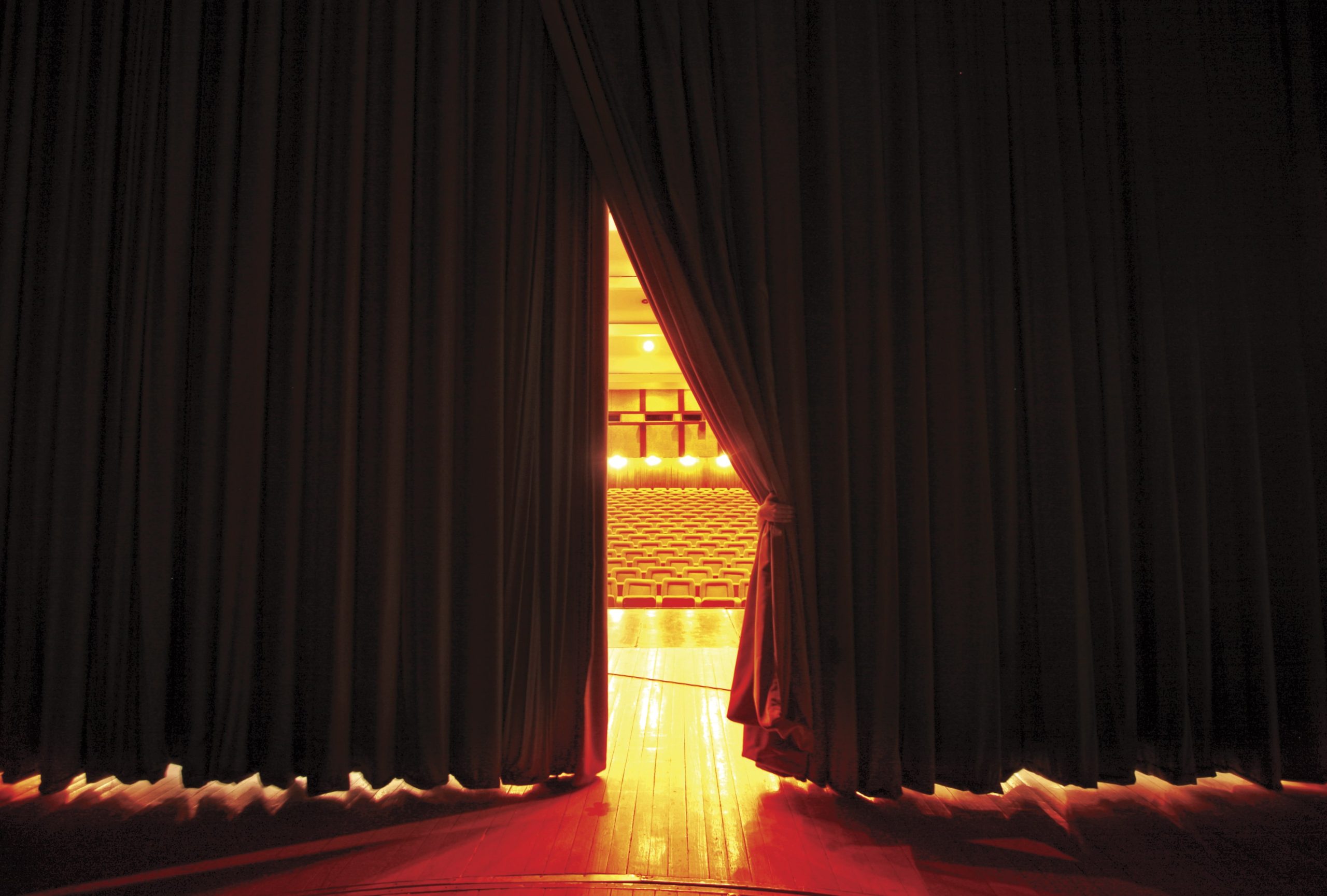 Vista de um palco com as cortinas se abrindo revelando bancos vazios em uma platéia.
